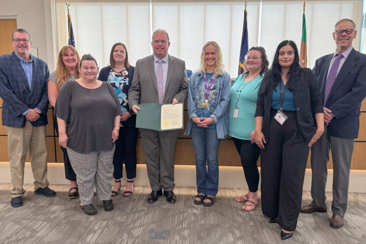 A group of business professionals pose for a photo. A man in the center holds open a proclamation to display.