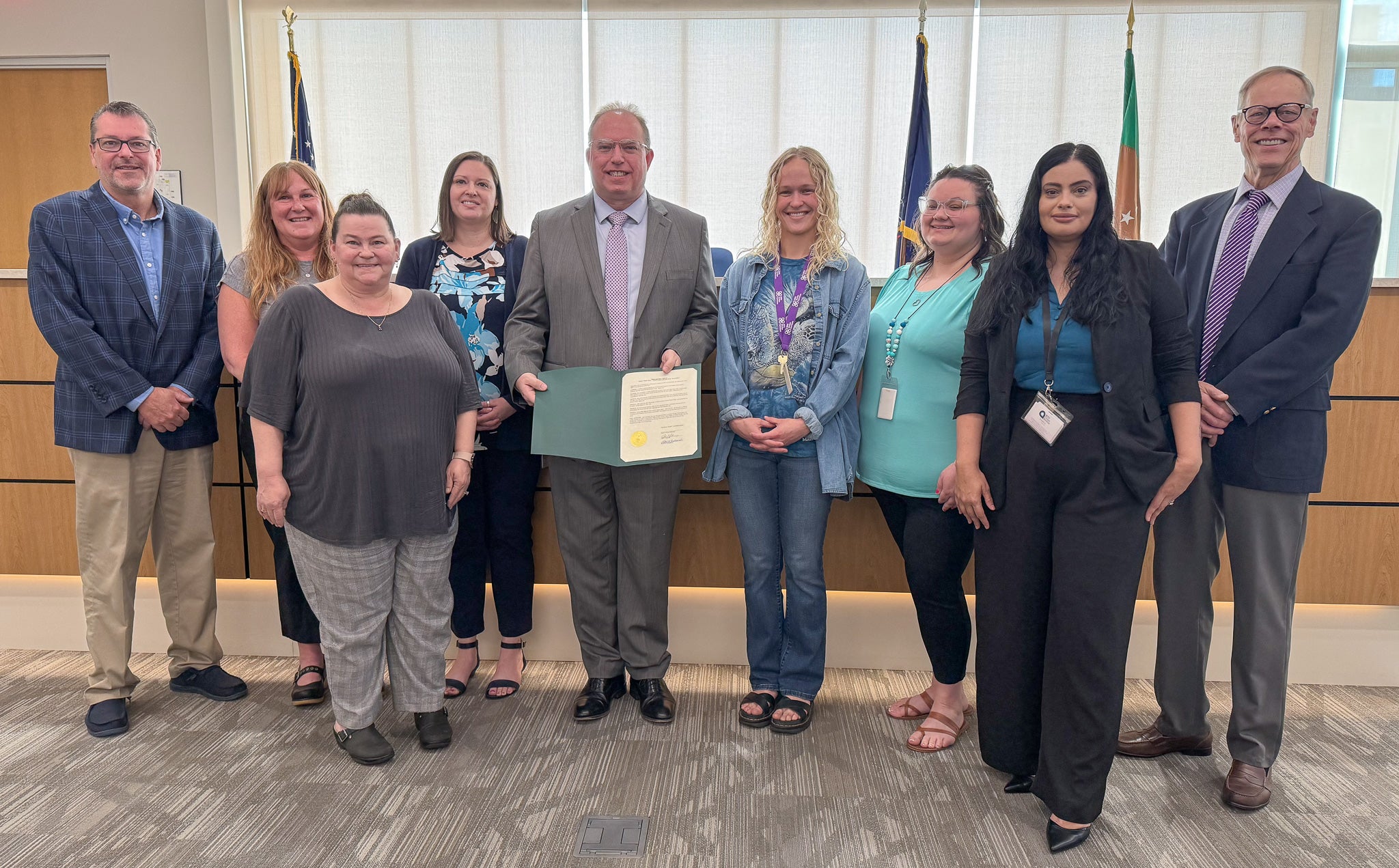 A group of business professionals pose for a photo. A man in the center holds open a proclamation to display.